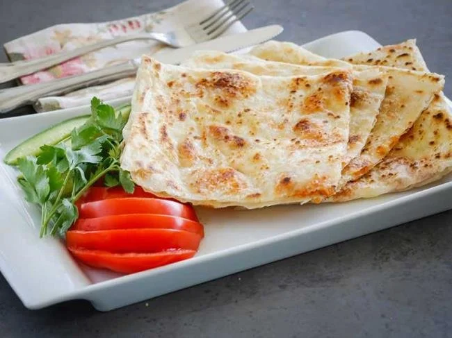 İnce dilimlenmiş naan ekmeği, yanında domates, salatalık ve taze kişniş ile servis edilen bir tabakta yer alıyor.