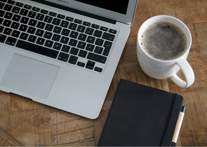 A laptop, a notebook with a pen, a cup of coffee, and a smartphone on a wooden table.
