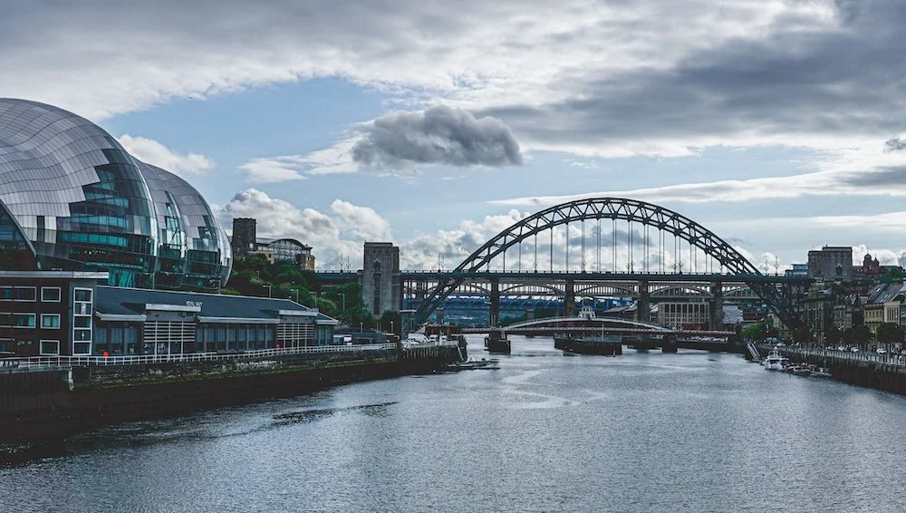 Cityscape with a river, a modern dome-shaped building, a bridge, and cloudy sky in the background.