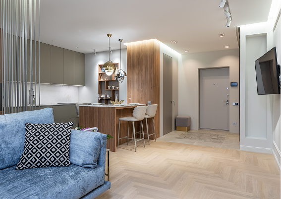 Modern living room with a blue sofa, kitchen with bar stools, built-in wooden shelving, and a mounted flat screen TV.