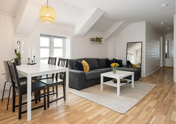 Living room with a gray sofa, white coffee table with a vase of flowers, wall mirror, dining table with six black chairs, window, and wooden floors.