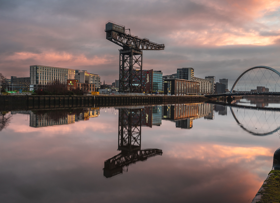 City skyline at dusk reflected in calm river, featuring a large industrial crane, modern buildings, and a bridge with an arch.