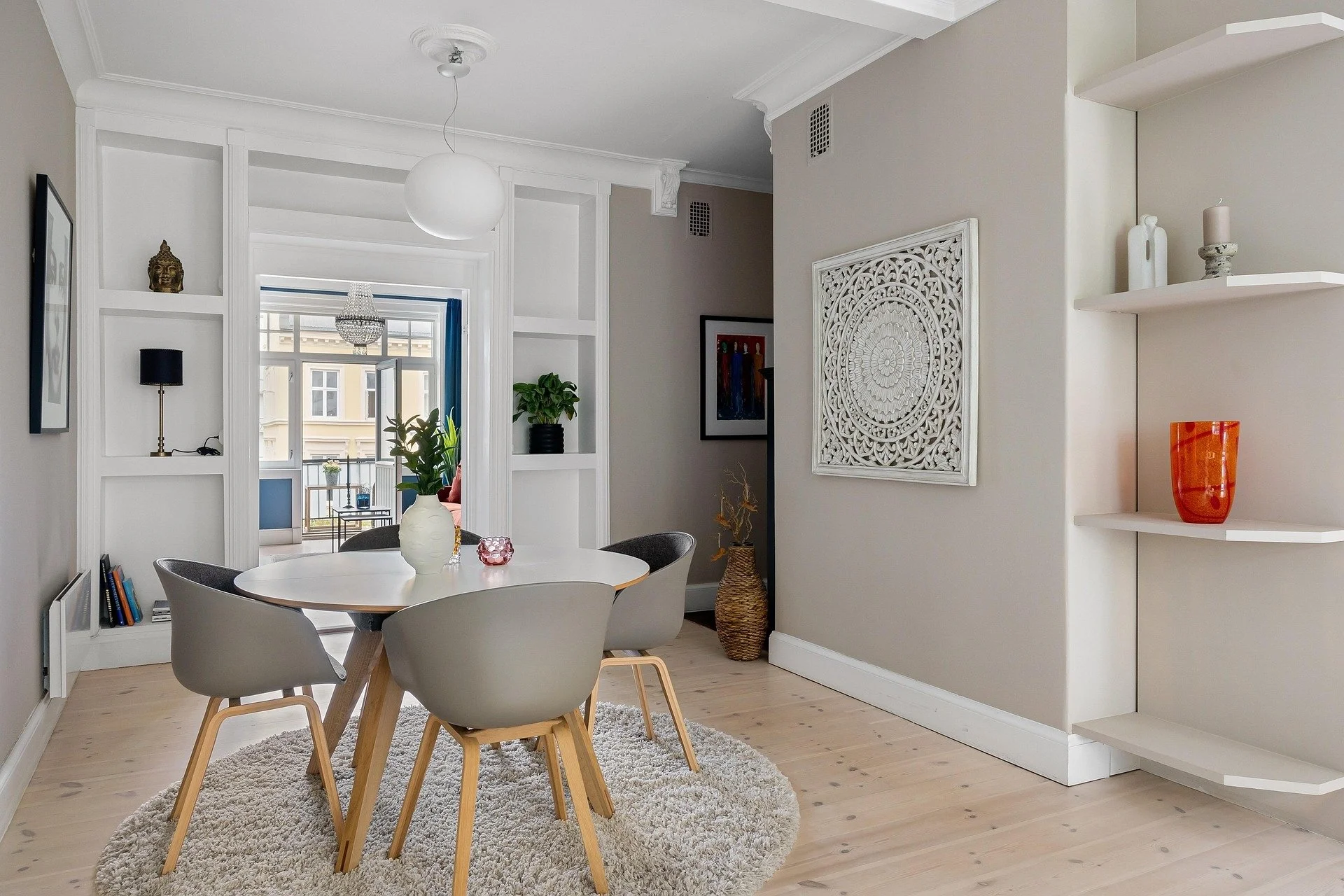 Interior of a modern dining room with a round white table, four gray chairs with wooden legs, a fluffy beige rug, and large windows with a balcony view. Decor includes a white carved wall panel, shelves with vases, a potted plant, and a hanging light fixture.