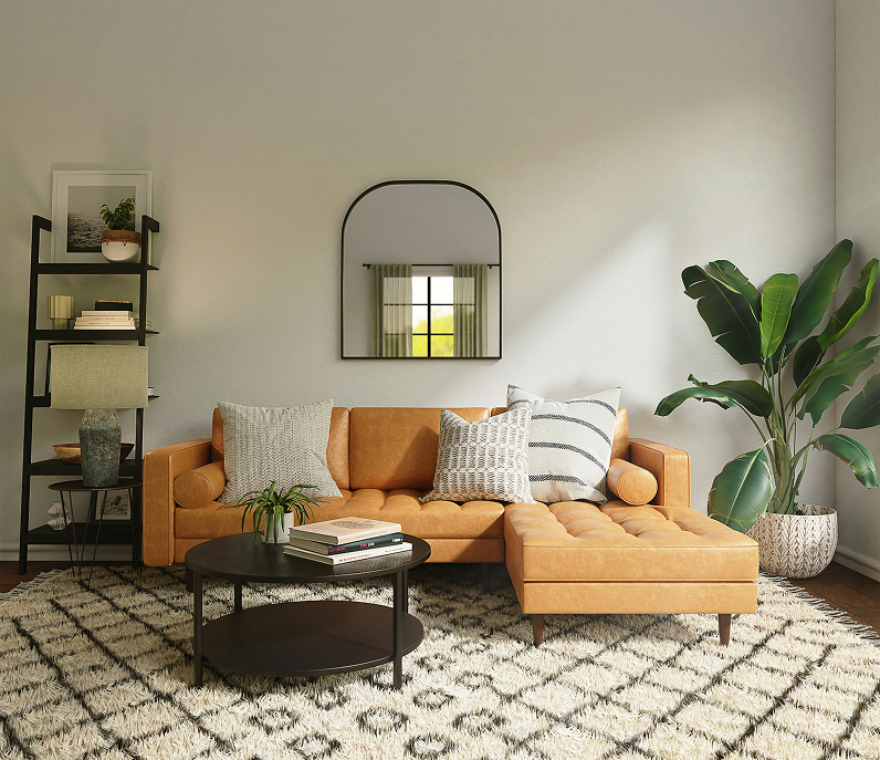 A cozy living room with a tan leather sectional sofa decorated with throw pillows, a black round coffee table with books and a small plant, a black bookshelf with decorative items, a large potted plant, a mirror on the wall, and a patterned rug.