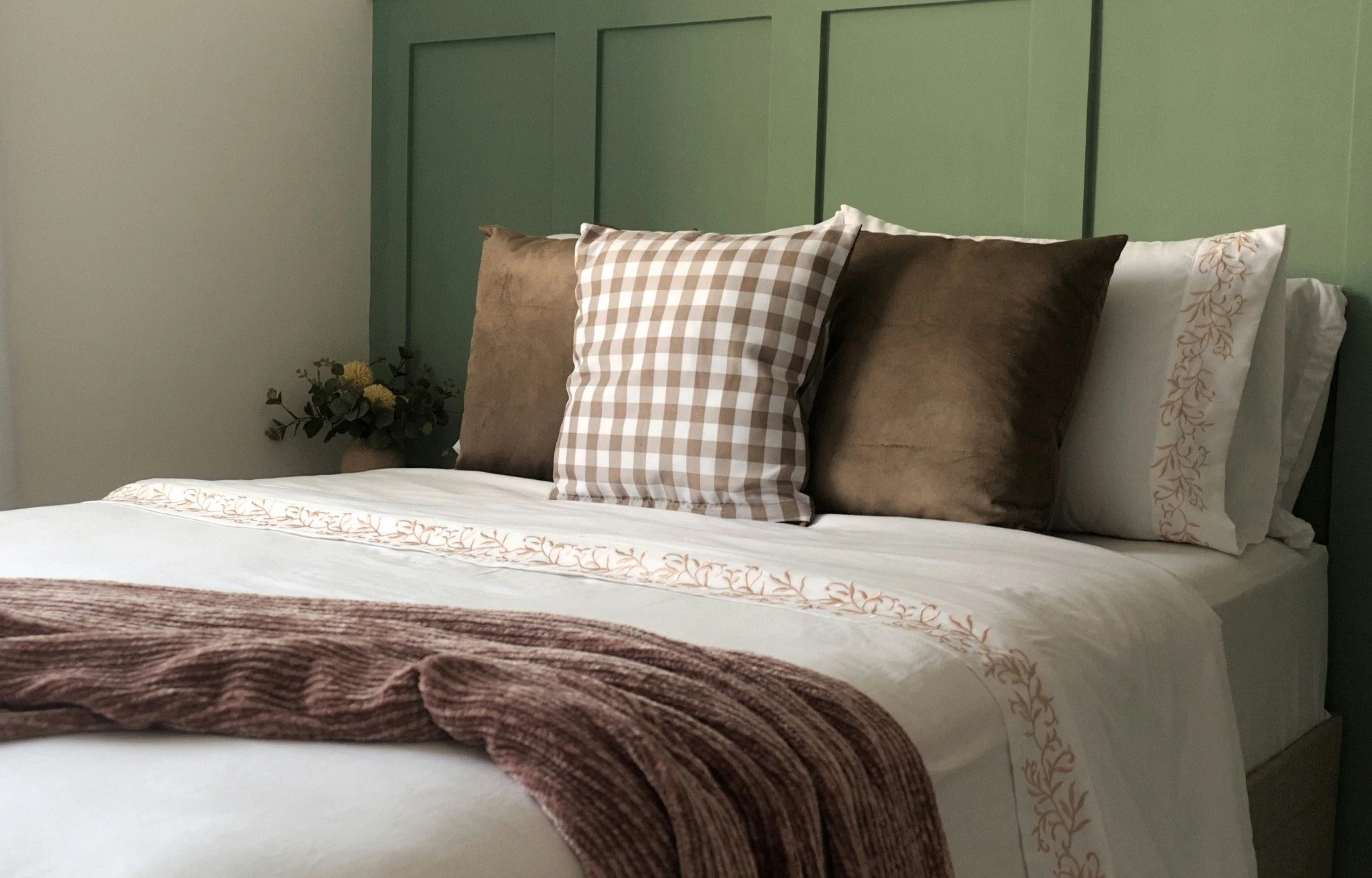 A neatly made bed with white sheets, a floral embroidered pillowcase, and a row of assorted pillows including beige and checkered patterns, accompanied by a small floral arrangement on the bedside table.
