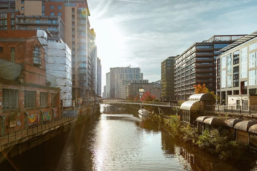 A cityscape with high-rise buildings along a river under a partly cloudy sky, with the sun shining behind the buildings.