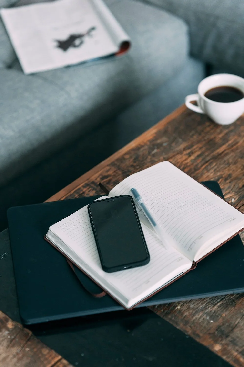 Open notebook with lined pages, a pen, a smartphone, a closed black folder, and a coffee cup on a wooden table. In the background, a gray sofa with a magazine on it.