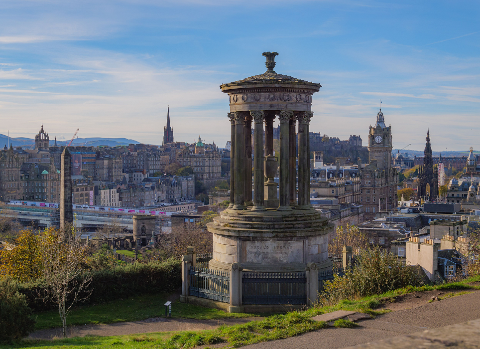 A stone monument with a cylindrical colonnade on a hill overlooking a cityscape with historic buildings and clock towers, under a partly cloudy blue sky.