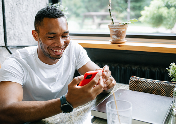 A man smiling and looking at his phone while sitting at a table in a cafe.