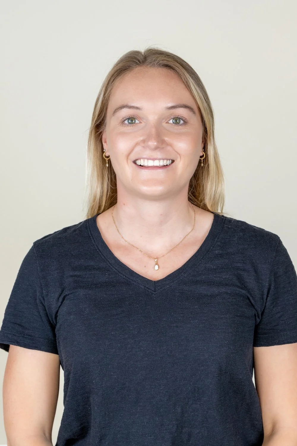Portrait of a smiling woman with blonde hair, green eyes, wearing a black t-shirt and gold jewelry against a plain beige background.