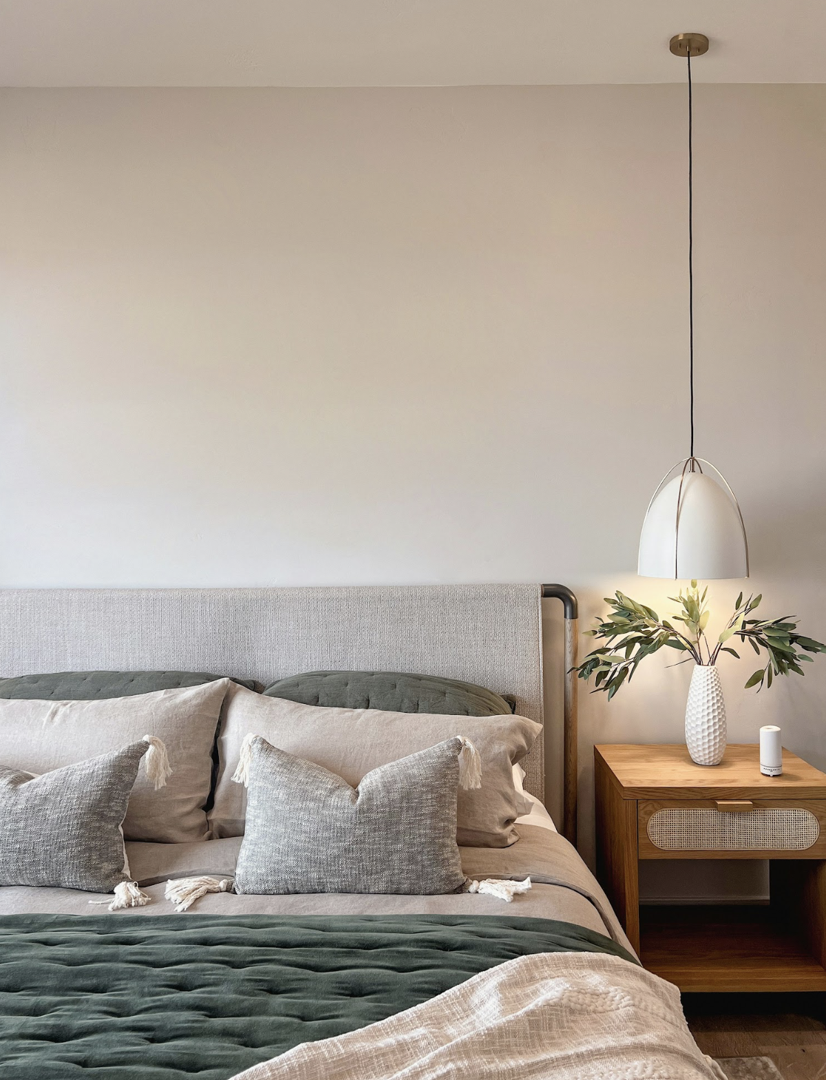 A neatly made bed with beige and green pillows, a textured beige headboard, and a wooden nightstand with a plant, white diffuser, and a white hanging pendant light in a minimalistic bedroom.