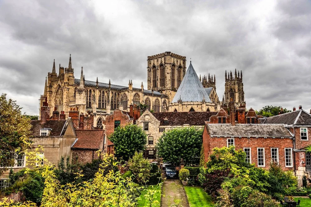 View of a historic Gothic cathedral with tall spires and ornate architecture, situated behind a neighborhood of residential houses with brick and stone exteriors, surrounded by lush greenery and trees under a cloudy sky.