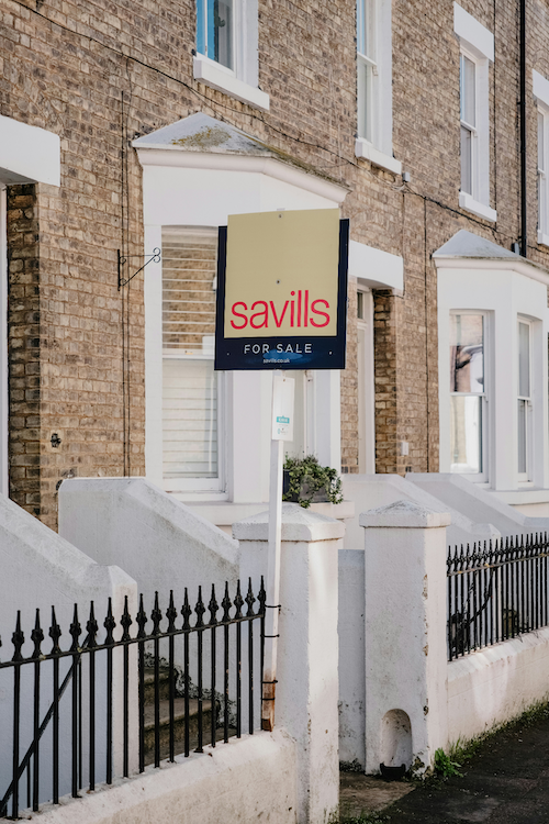 A for sale sign in front of a row of brick townhouses with white steps and black iron fencing.