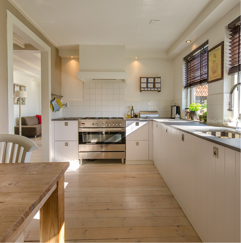 Bright kitchen with white cabinets, a stainless steel oven, and a window with brown blinds. Potted plants and kitchen items are on the countertop.