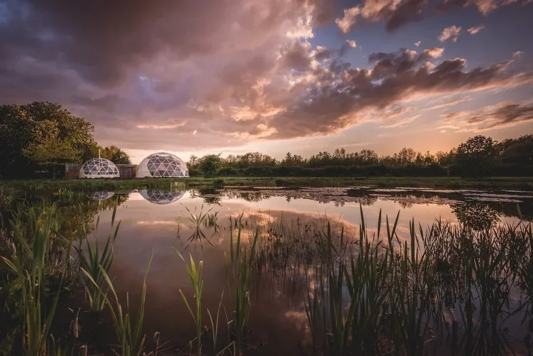 Two geodesic domes beside a pond reflecting a dramatic sunset sky.