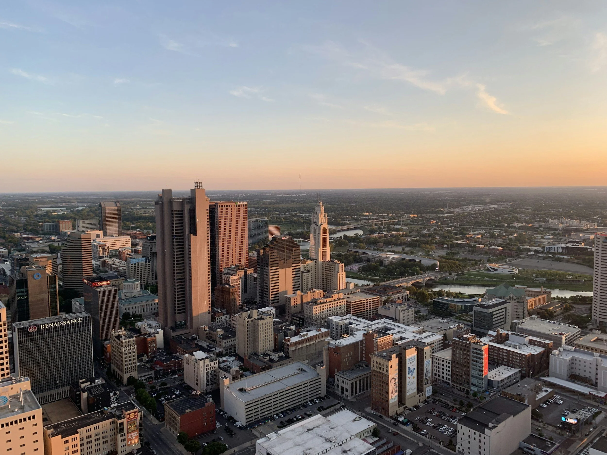 City skyline at sunset with tall buildings, a river, and a clear sky with some clouds.