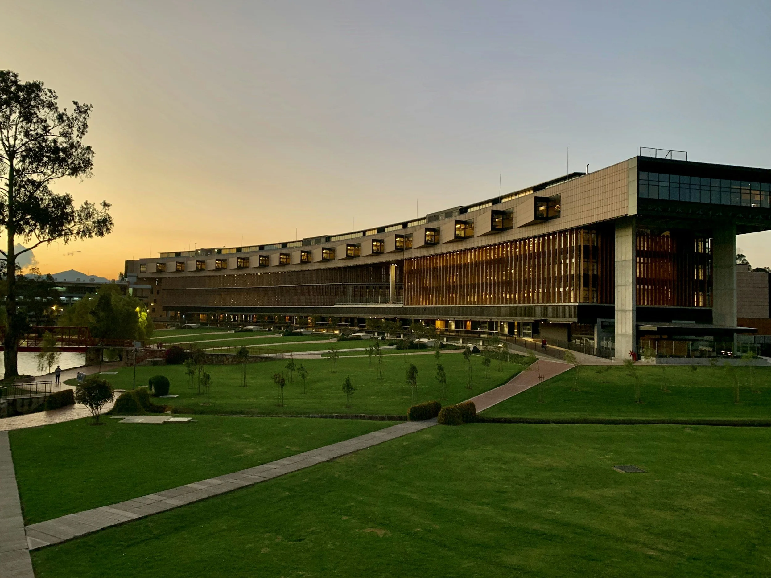 A modern building with large windows, a curved design, and illuminated interior spaces, set amidst a green landscaped area with pathways, trees, and a pond, under a clear evening sky.