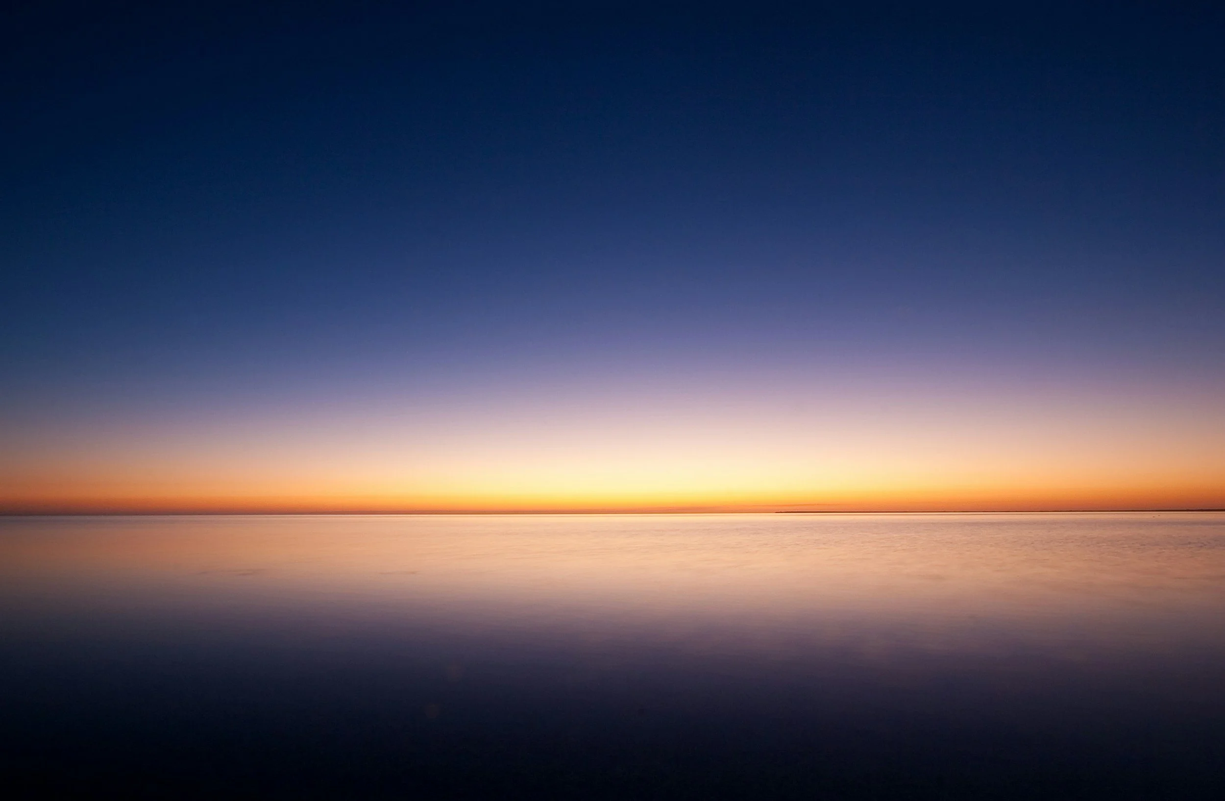 A tranquil scene of a sunset over a calm body of water, with the sky transitioning from orange near the horizon to deep blue higher up.