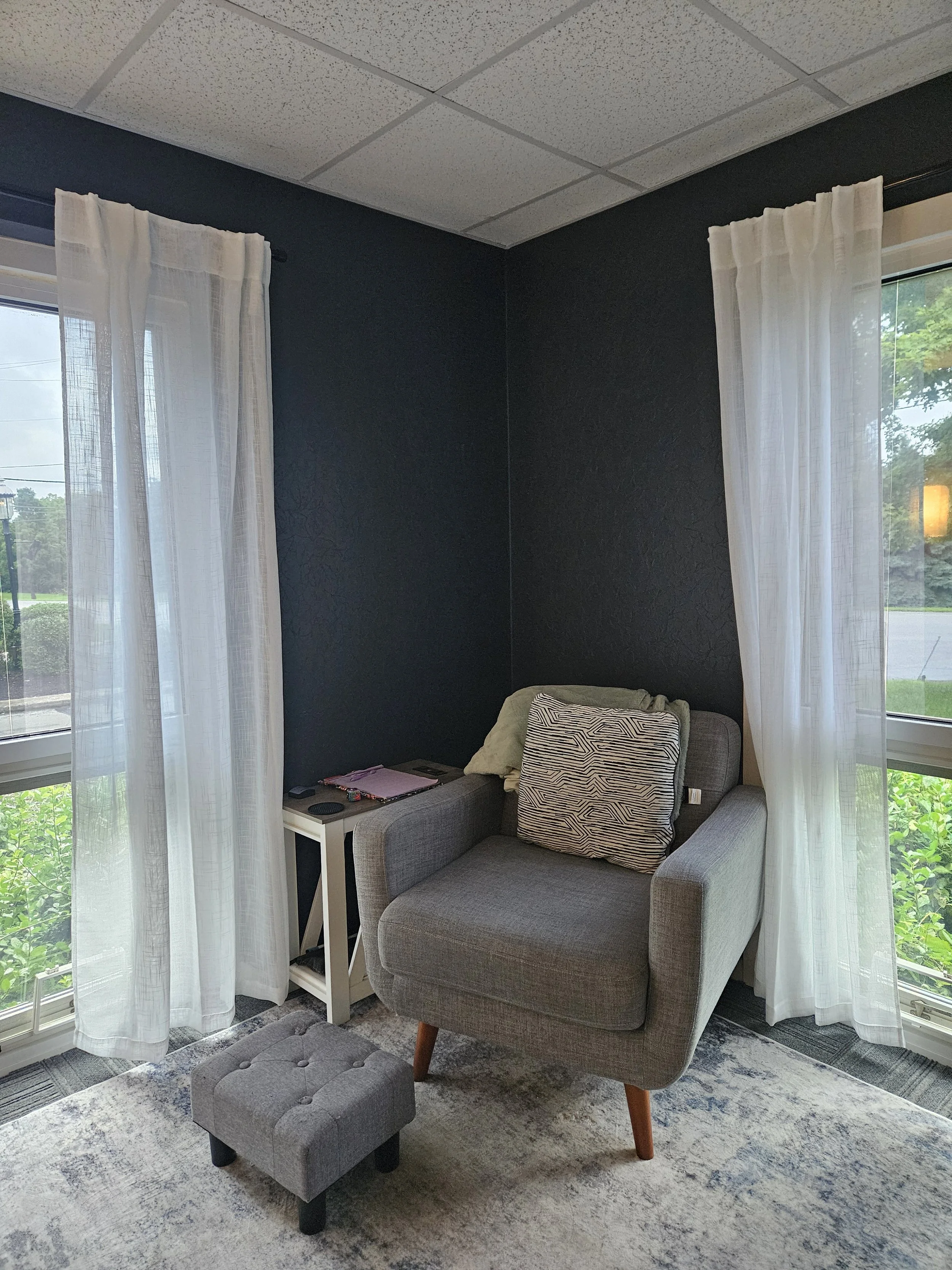 A cozy corner in the therapy room with a gray armchair and a matching small ottoman, next to a small white side table, surrounded by large windows with white curtains, and a view of green trees outside.