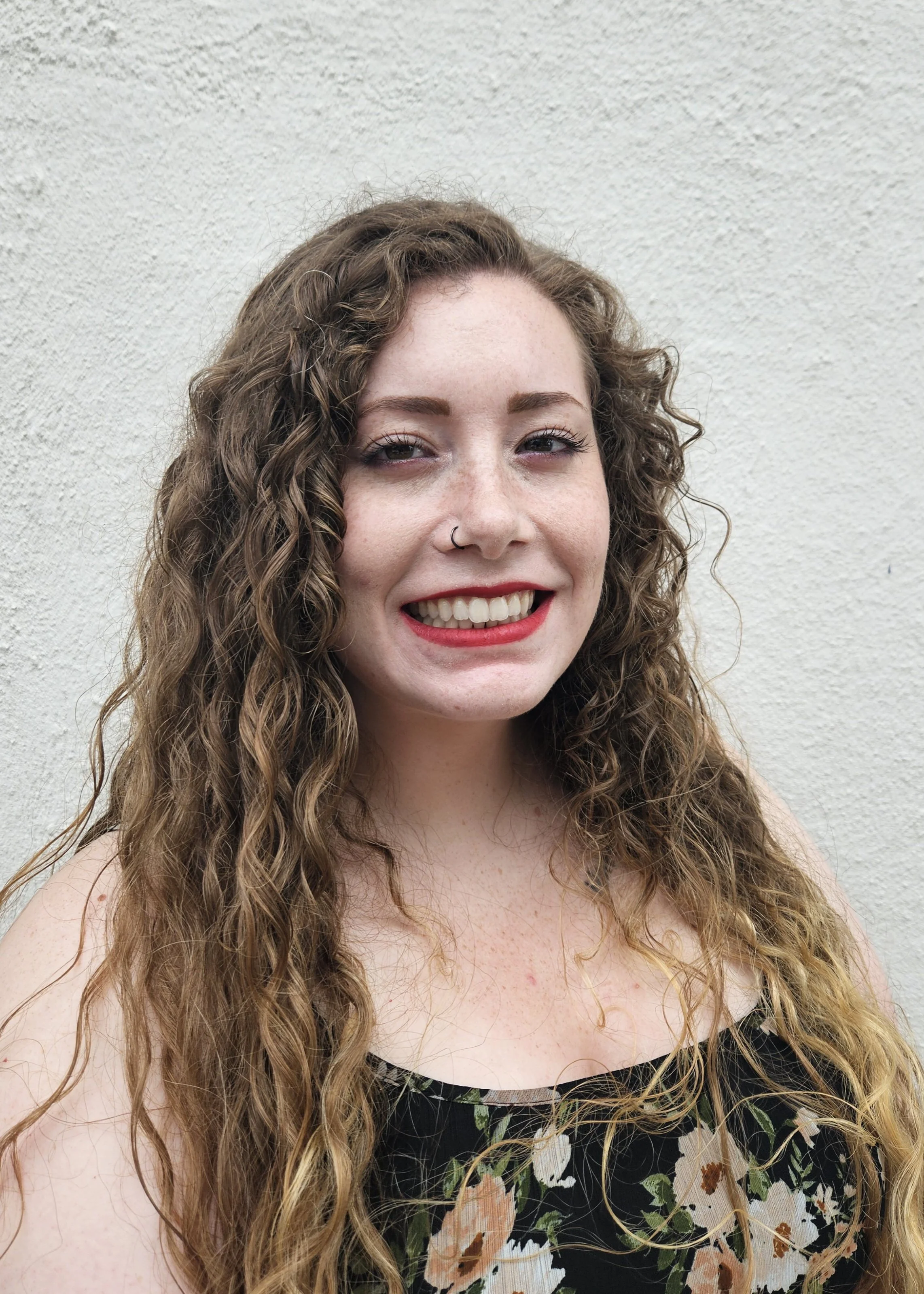 A young woman with long, curly brown hair smiling in front of a white textured wall, wearing a floral dress.