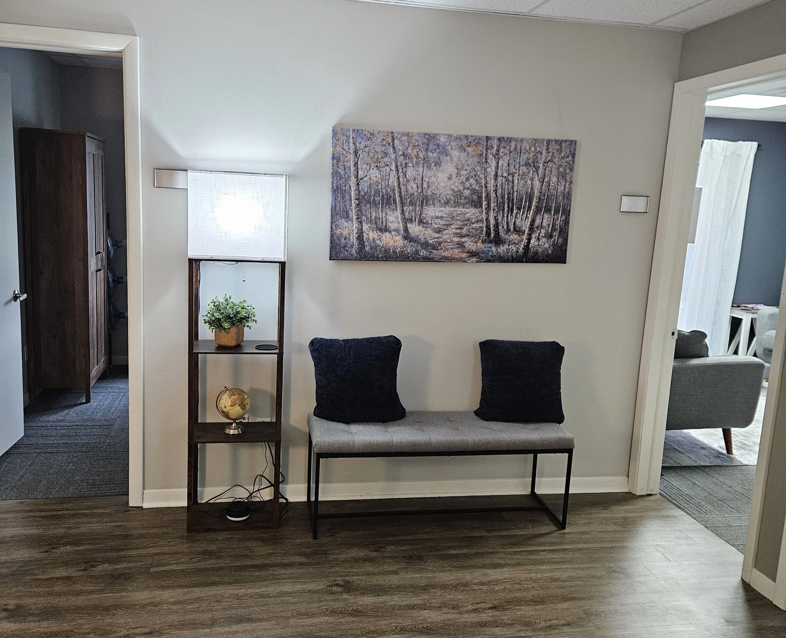 Interior view of a therapy waiting room with a wooden side table, a potted plant, a framed picture, a bench with two cushions, a large painting of a forest scene, and a globe on the floor. A doorway on the right shows a glimpse of the therapy room.