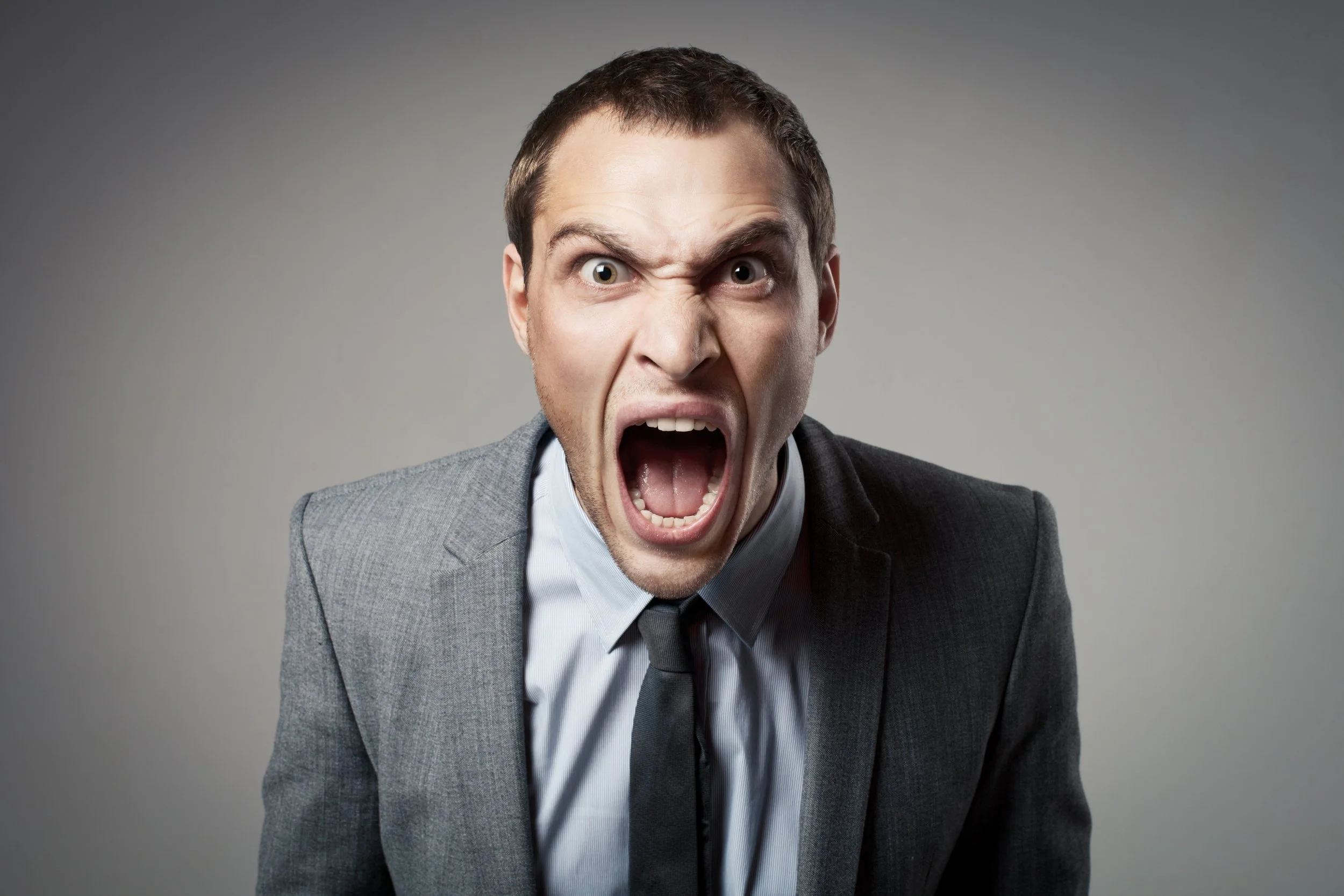 A man in a gray suit and tie with a white shirt is shouting with his mouth wide open, eyes wide, and eyebrows furrowed, expressing anger or frustration.