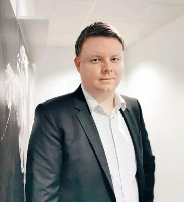 Junge männliche Person in einem schwarzen Anzug und weißem Hemd, steht vor einer weißen Wand.