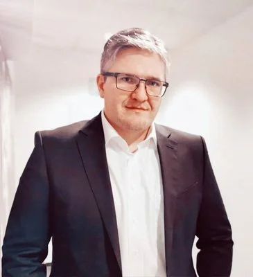 Mann in formellem Anzug und Brille auf einem weißen Hintergrund.
