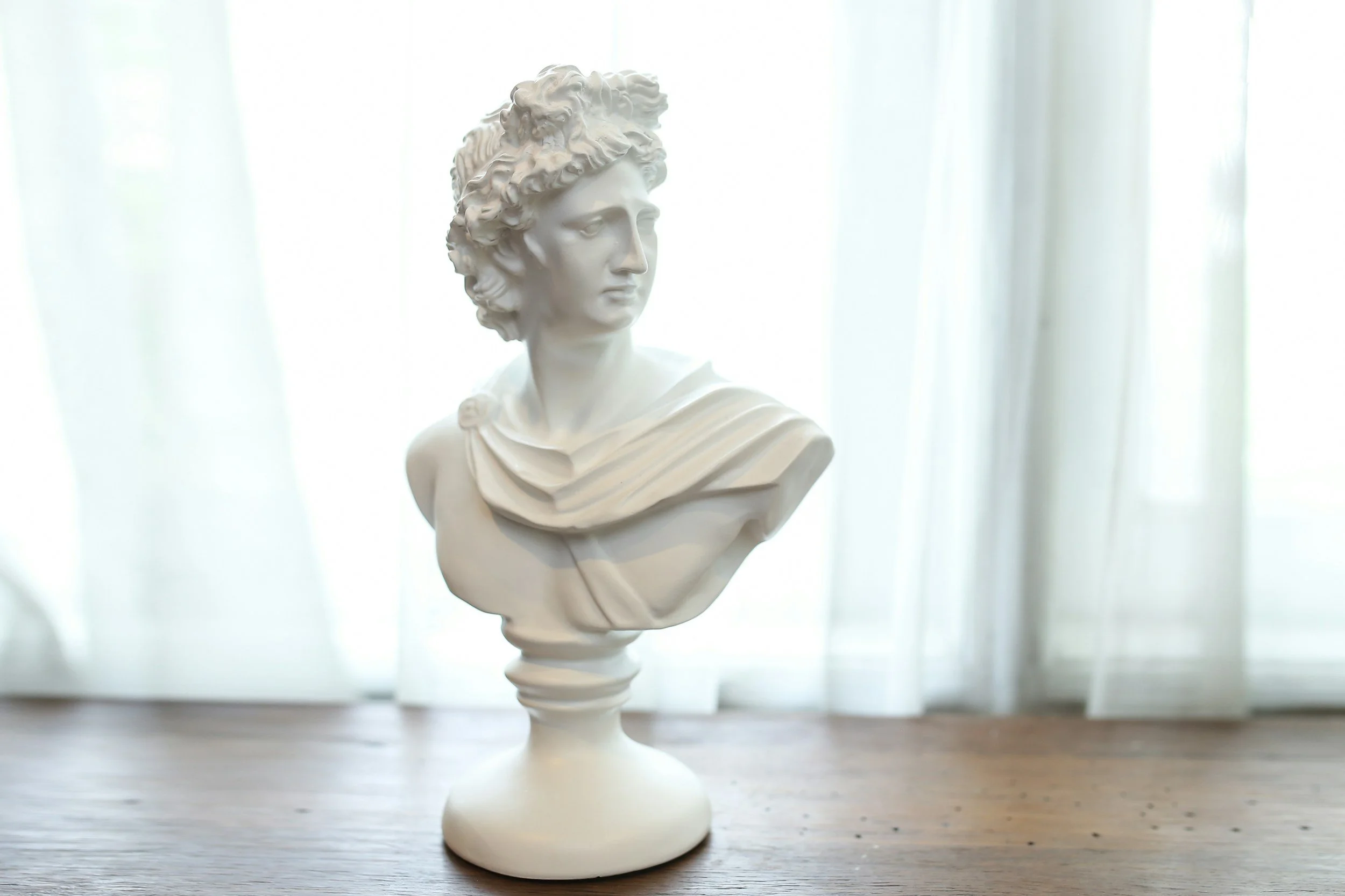 White bust sculpture of a woman's head and shoulders on a round base, placed on a wooden surface with a light curtain background.