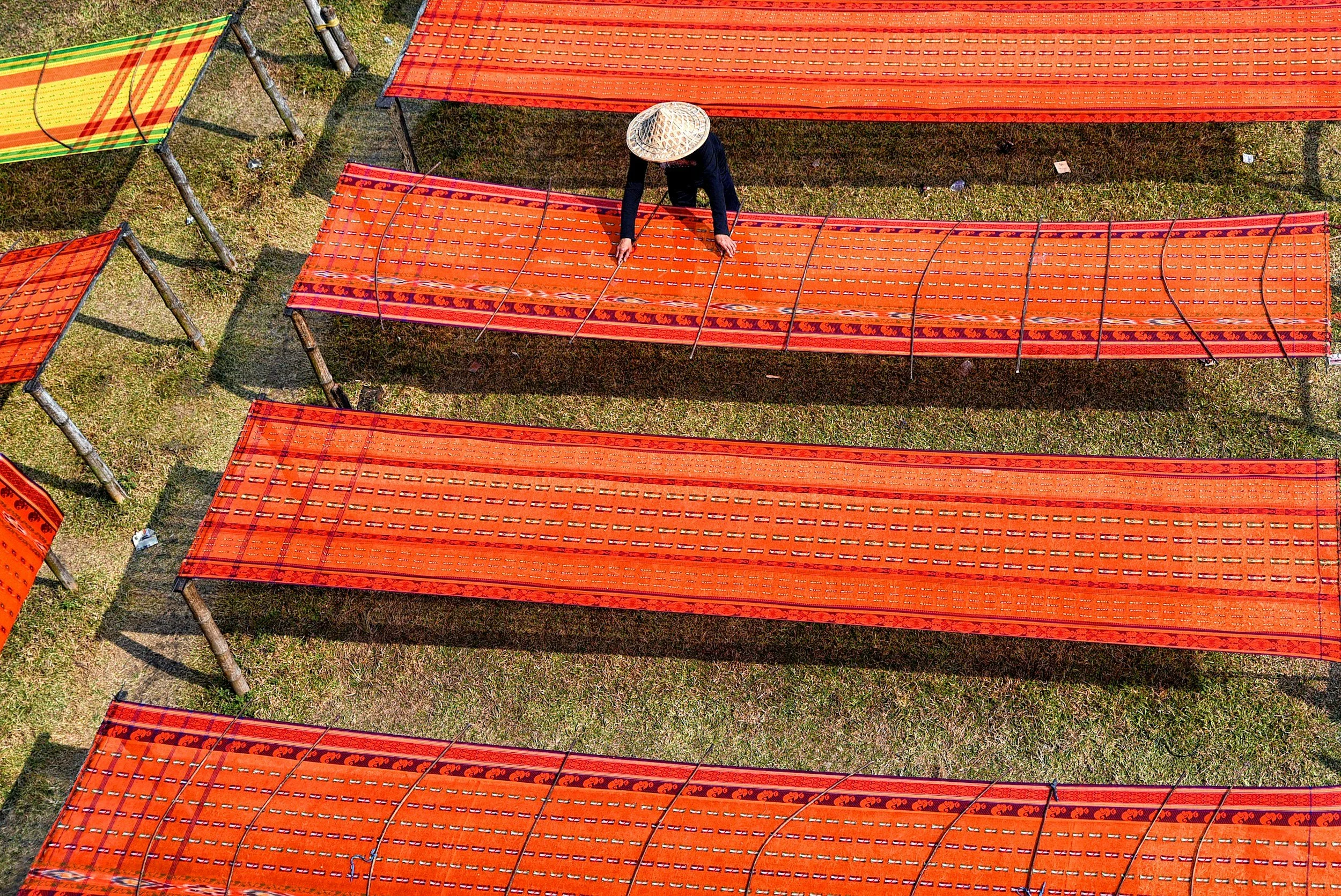 A person wearing a conical Asian hat watering orange plants arranged on raised beds outdoors on grassy ground.