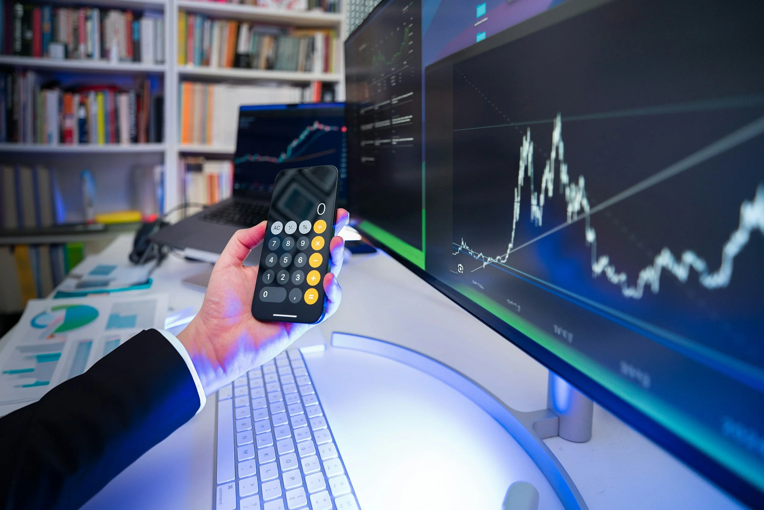 A person in a suit holding a calculator in front of computer screens displaying stock market graphs in an office setting.