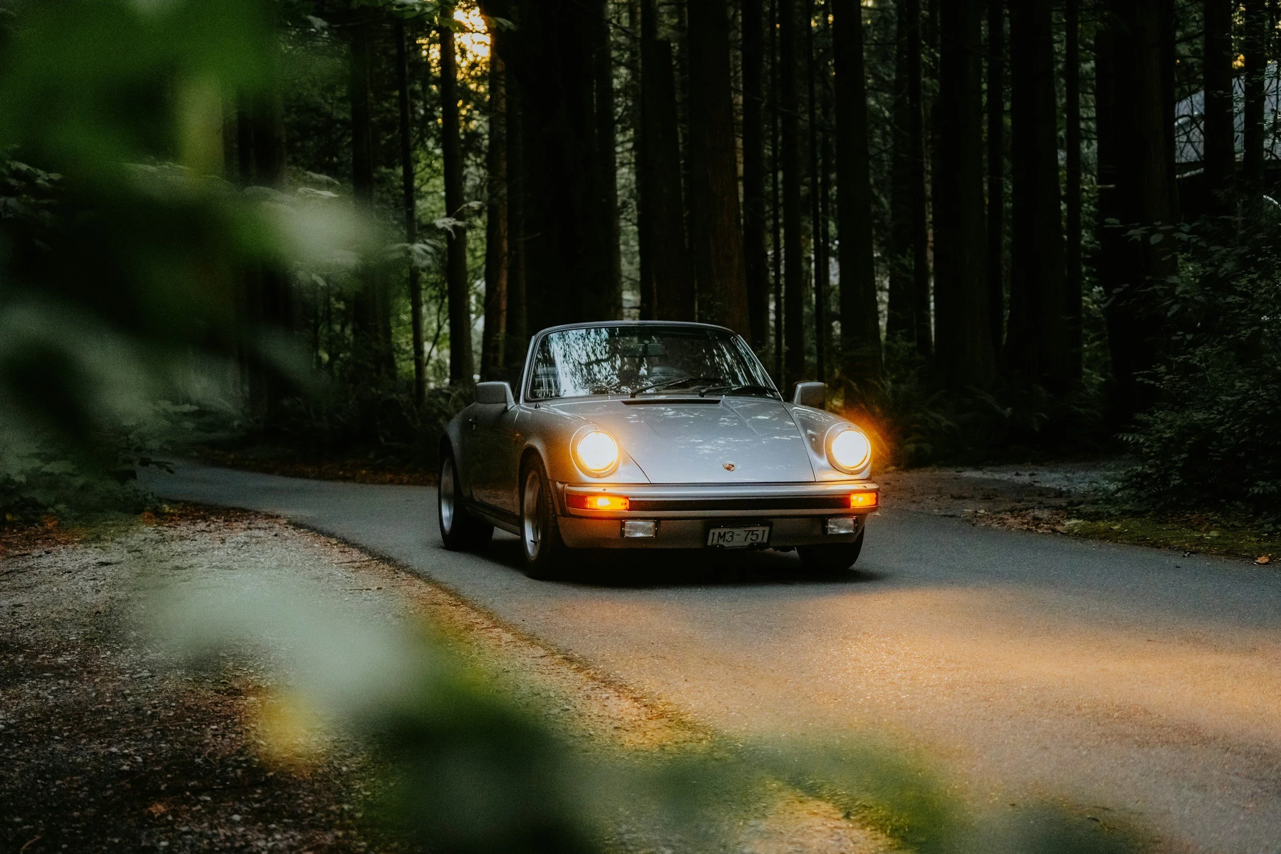 A gray Porsche with its headlights on driving on a narrow forest road during dusk, surrounded by tall trees.