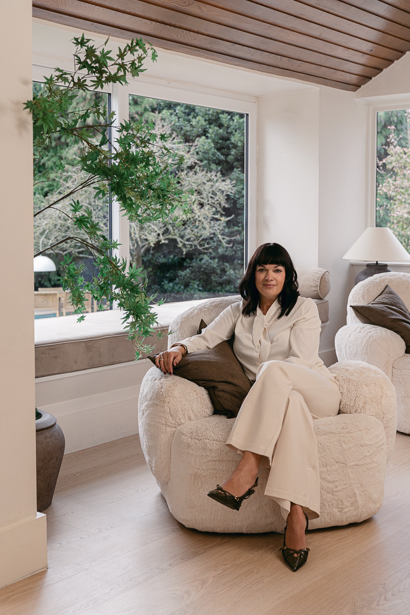 A woman sitting on a plush, white, fluffy armchair in a bright living room with large windows showing greenery outside. She is wearing a white suit and black high heels, with dark hair and bangs, and she is looking at the camera.
