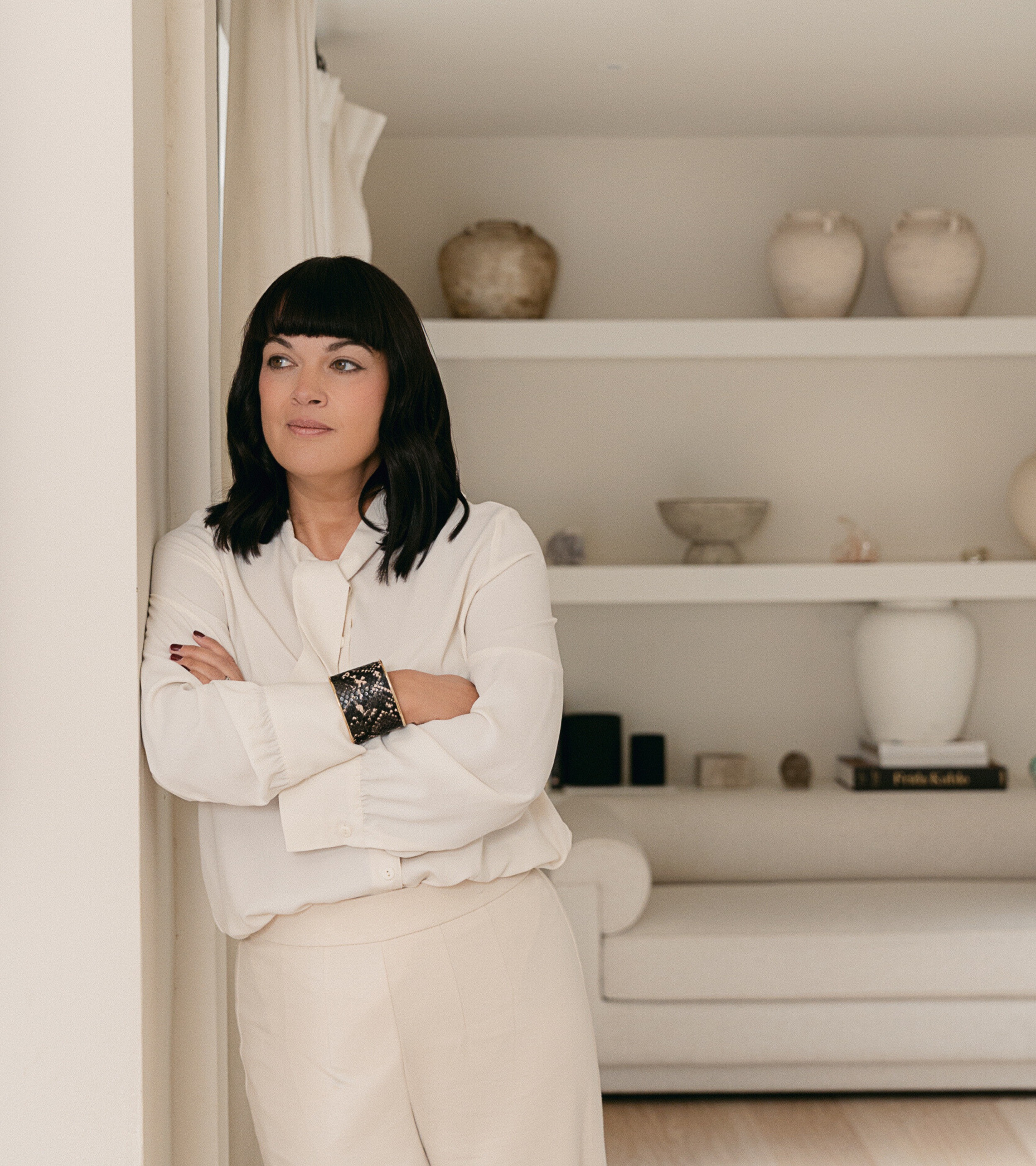 A woman with black hair and bangs wearing a white blouse and cream pants, standing in a room with shelves that have decorative vases and bowls.