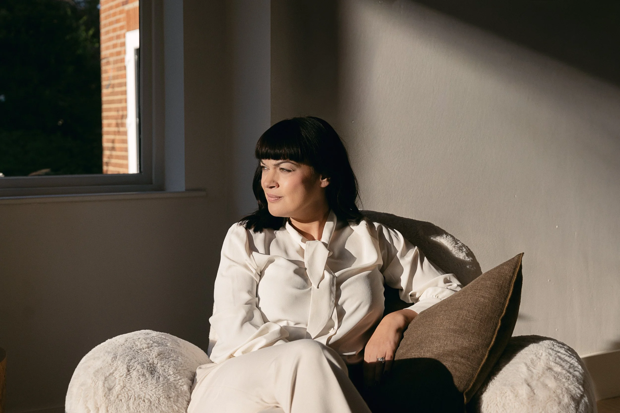 A woman with black hair and bangs sitting on a couch in a Sunlit room, looking outside through a window.