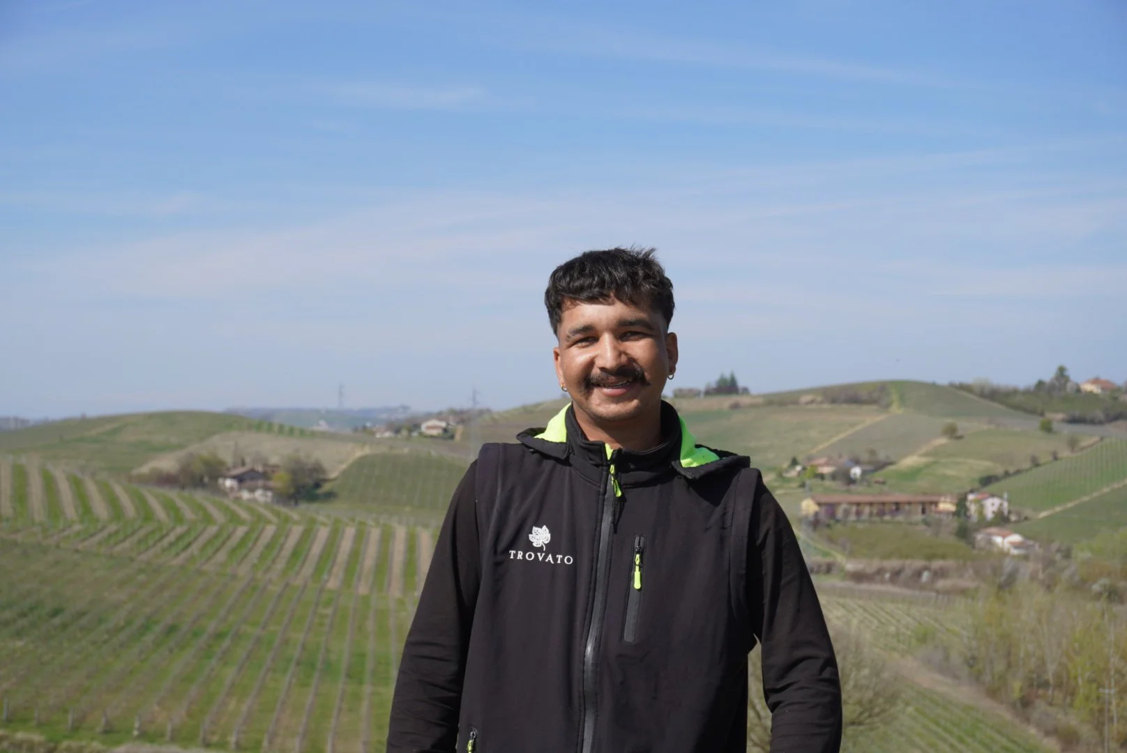 Uomo con barba e capelli ricci indossa giacca nera con marchio Trovato, sorridente, in campagna con colline e vigneti sotto un cielo sereno.