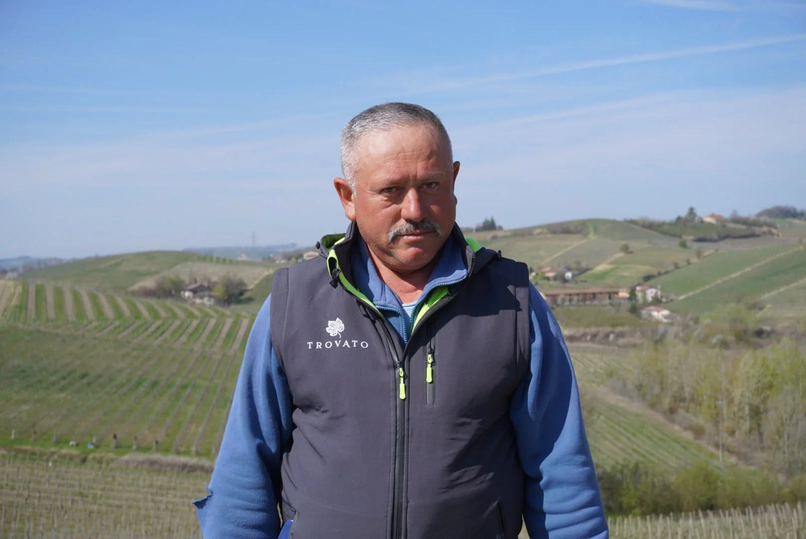 Uomo con cappotto blu e gilet nero che si trova in un paesaggio collinare con vigneti, sotto un cielo sereno.