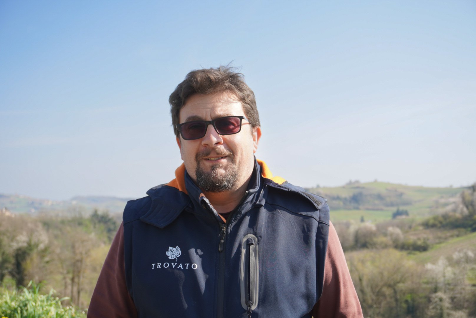 Uomo con occhiali da sole in abbigliamento casual all'aperto, con paesaggio collinare verde e cielo sereno sullo sfondo.
