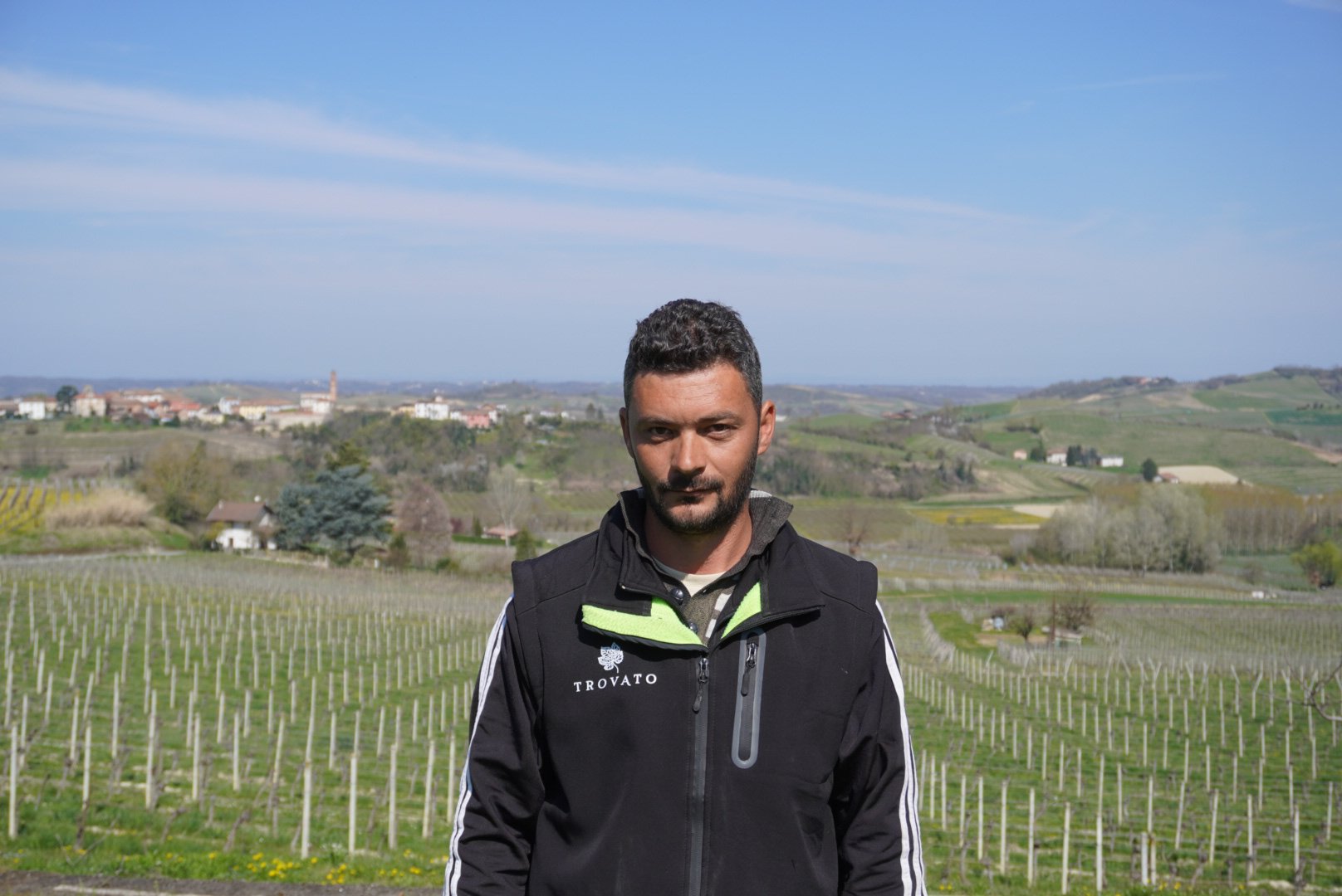 Uomo con giacca in piedi in un vigneto con paesaggi collinari sullo sfondo.