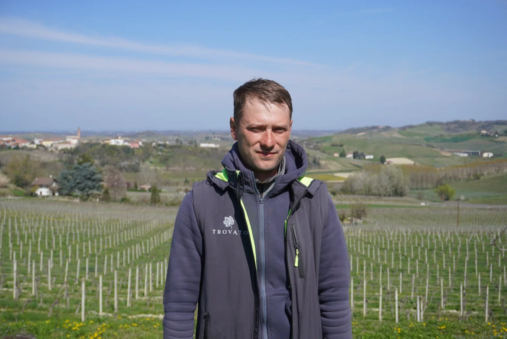 Uomo adulto con giacca in outdoor con paesaggio di campagna e vigneti sullo sfondo, cielo blu.