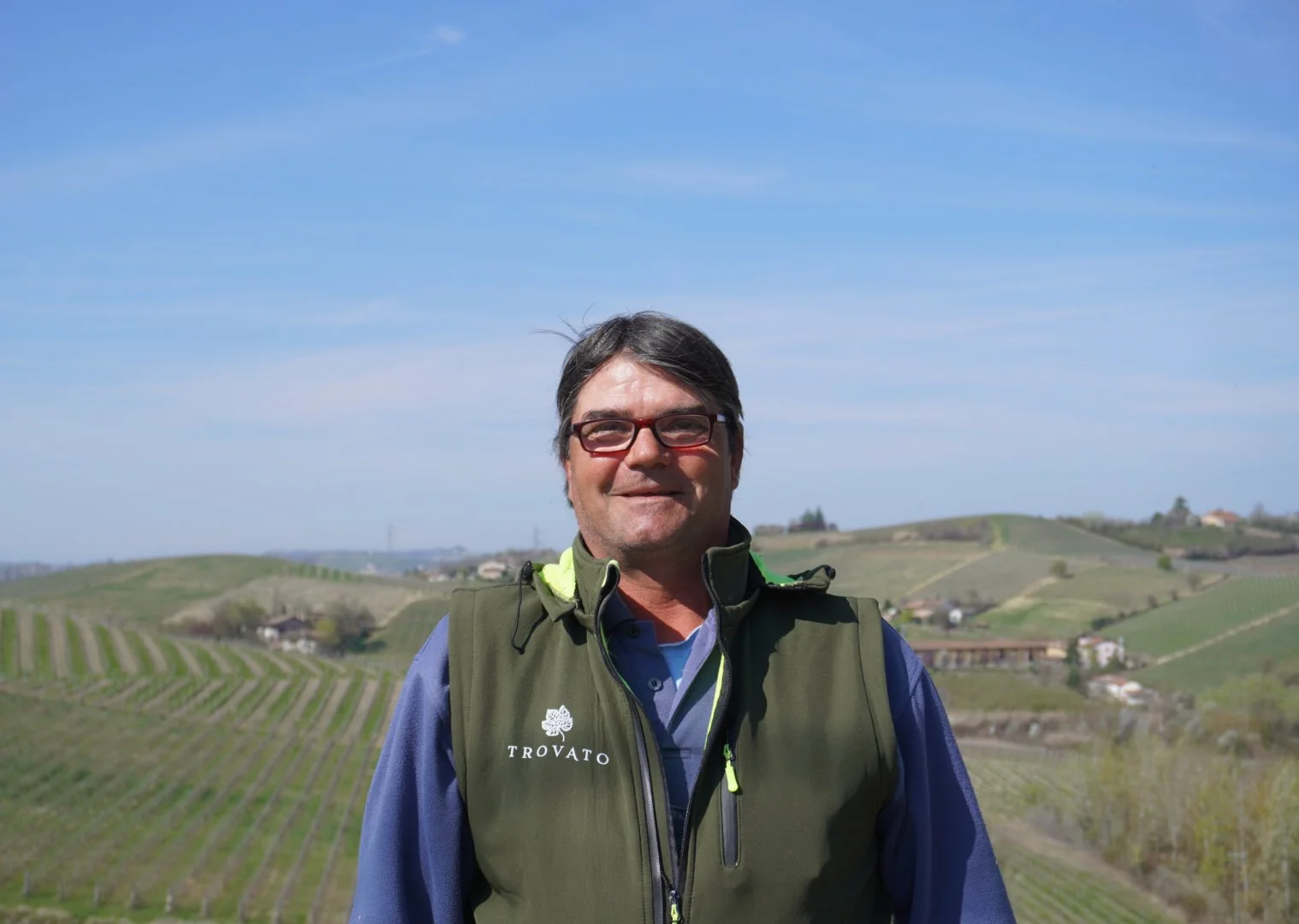 Uomo con occhiali e giacca a vento in piedi in un paesaggio collinare con vigneti sotto un cielo sereno.