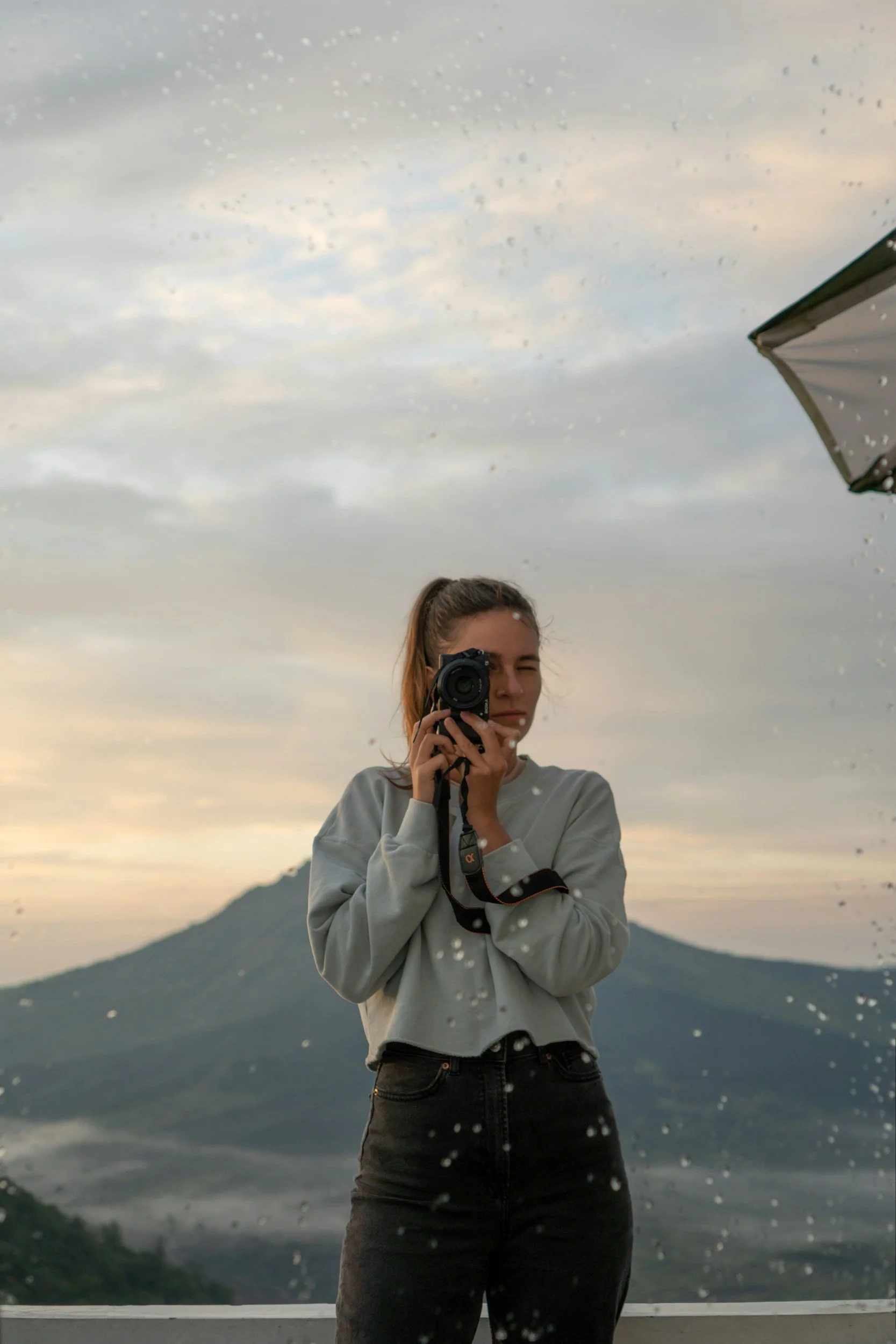 A young woman with reddish hair taking a photo in a rainy outdoor setting, with mountains and a cloudy sky in the background, seen through a rain-covered window.