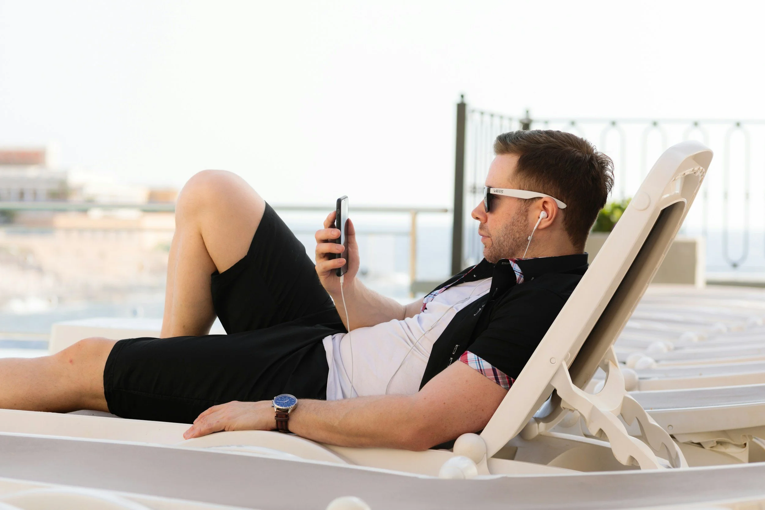 A man lounging on a beach chair, wearing sunglasses and headphones, looking at his phone with a backdrop of a bright outdoor setting.