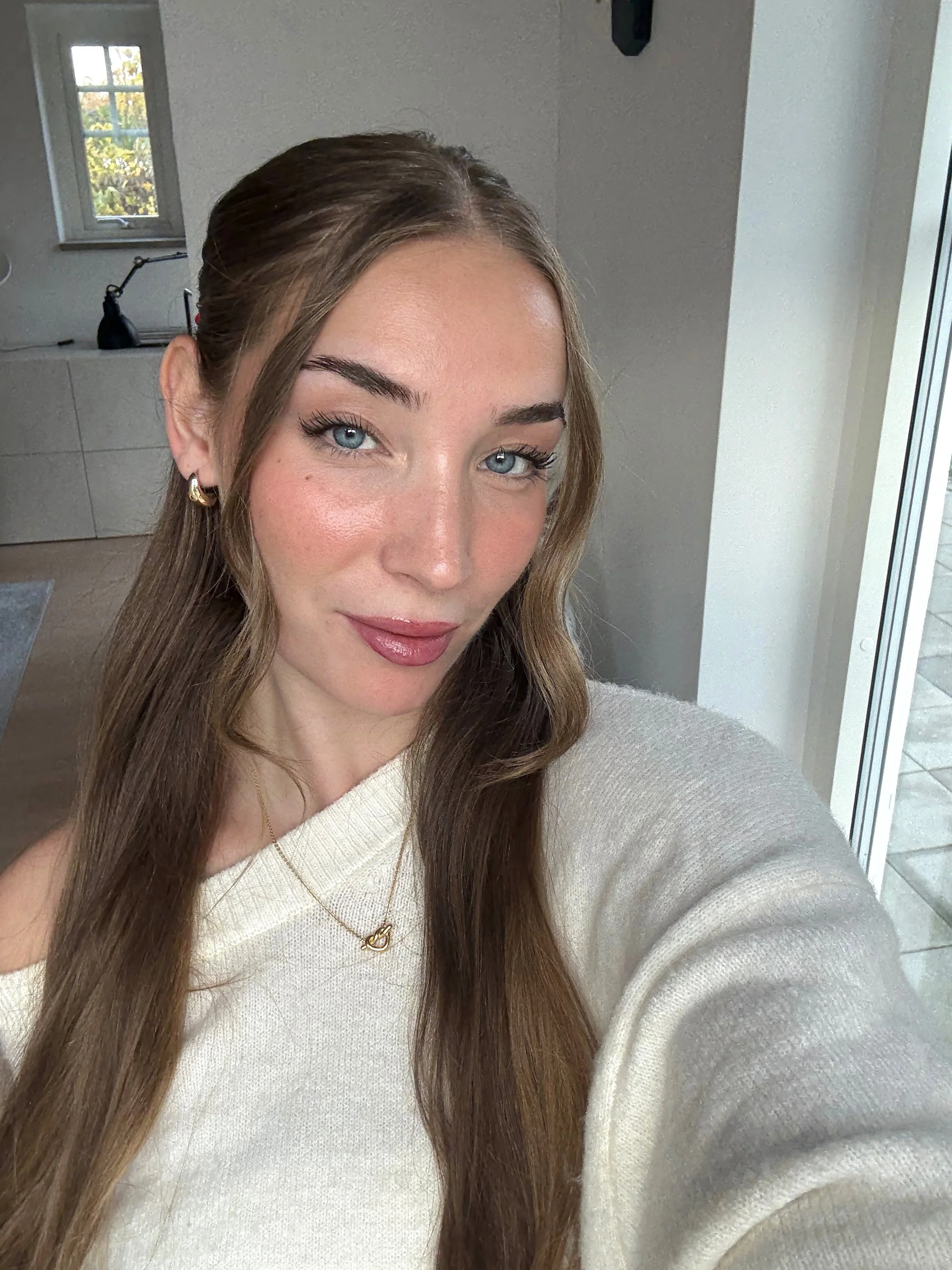 Close-up of a young woman with long brown hair and blue eyes, wearing gold earrings and a white sweater, smiling slightly at the camera. She is indoors near a window with a view of trees outside.