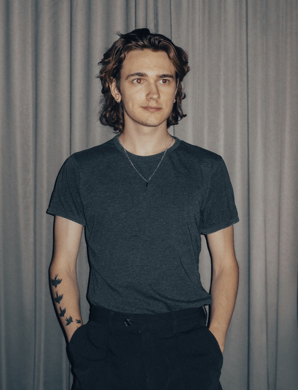 A young man with long, wavy brown hair stands in front of gray curtains, wearing a dark gray t-shirt and black pants, with tattoos of birds on his left arm and jewelry including earrings and a necklace.