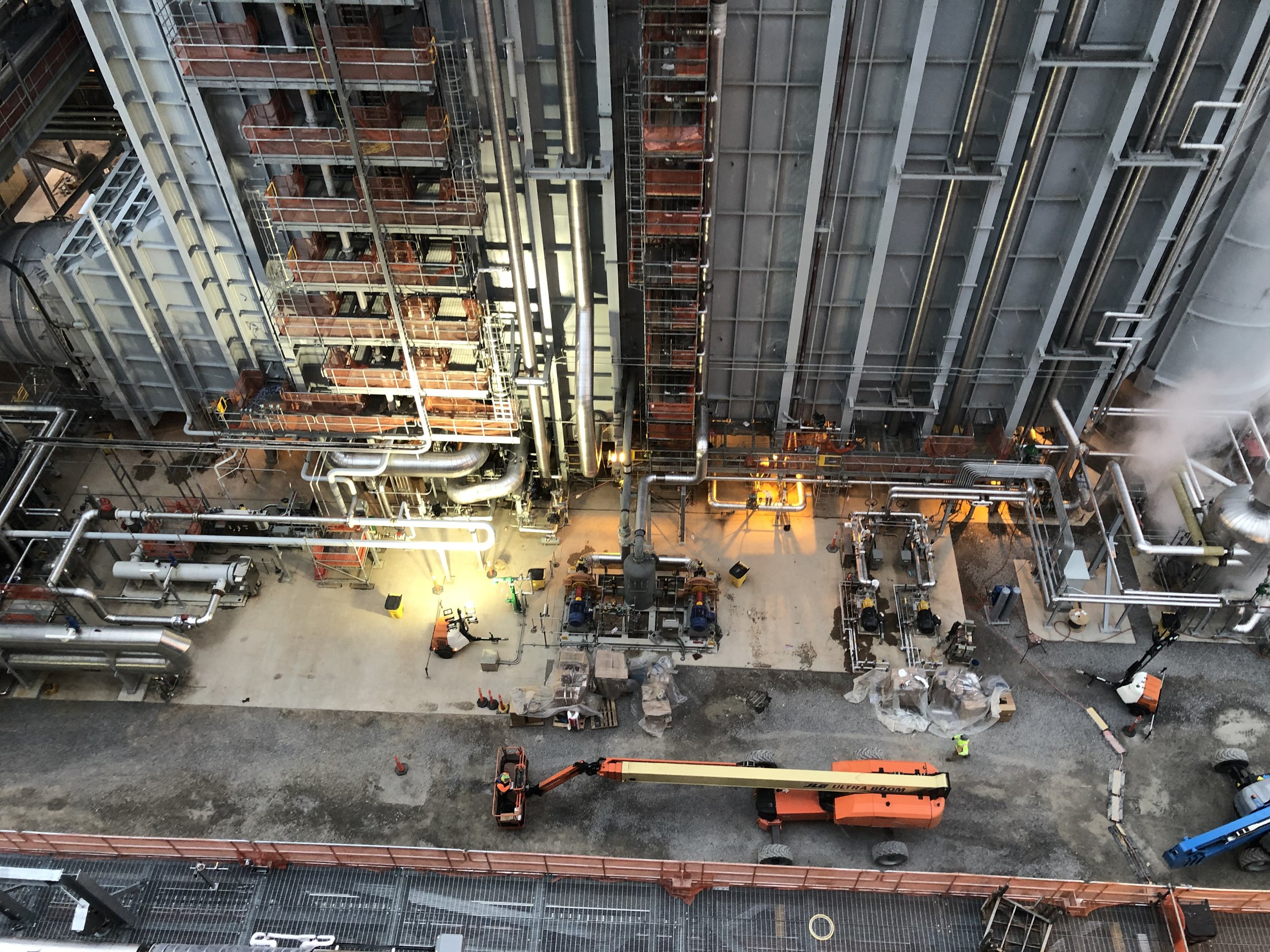 Construction workers and equipment on a building site with scaffolding, pipes, and safety cones.
