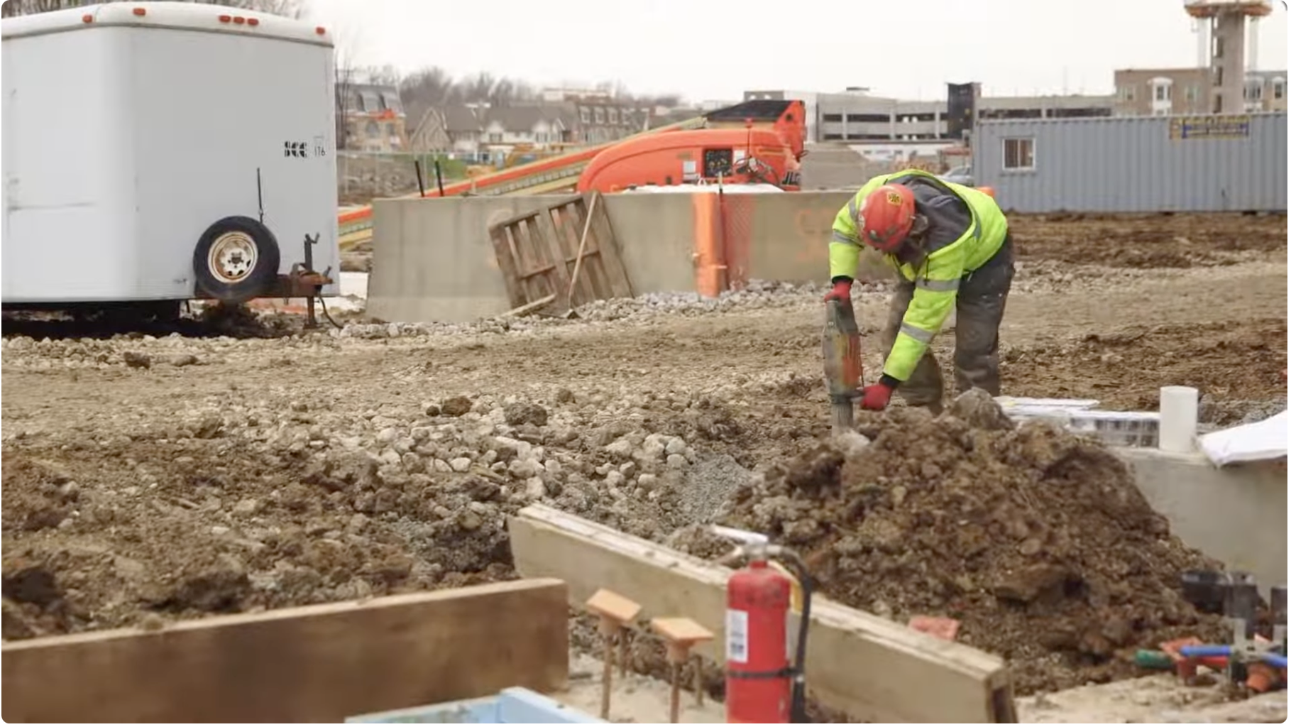 A construction worker wearing a yellow safety jacket, red helmet, and gloves is working with heavy equipment on a dirt construction site. There is construction equipment, building materials, and a container in the background.