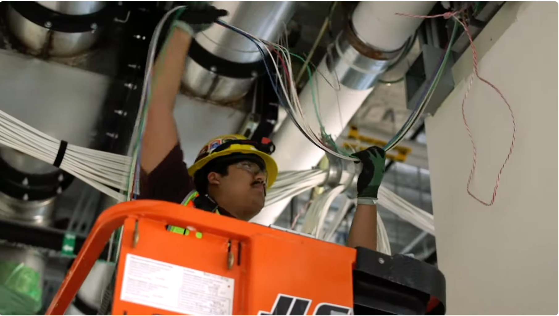 A worker wearing safety glasses, a yellow helmet, and a harness is working on electrical wiring and equipment inside an industrial or utility facility. The worker is using a tool to connect or repair wires near large metal pipes and cables.