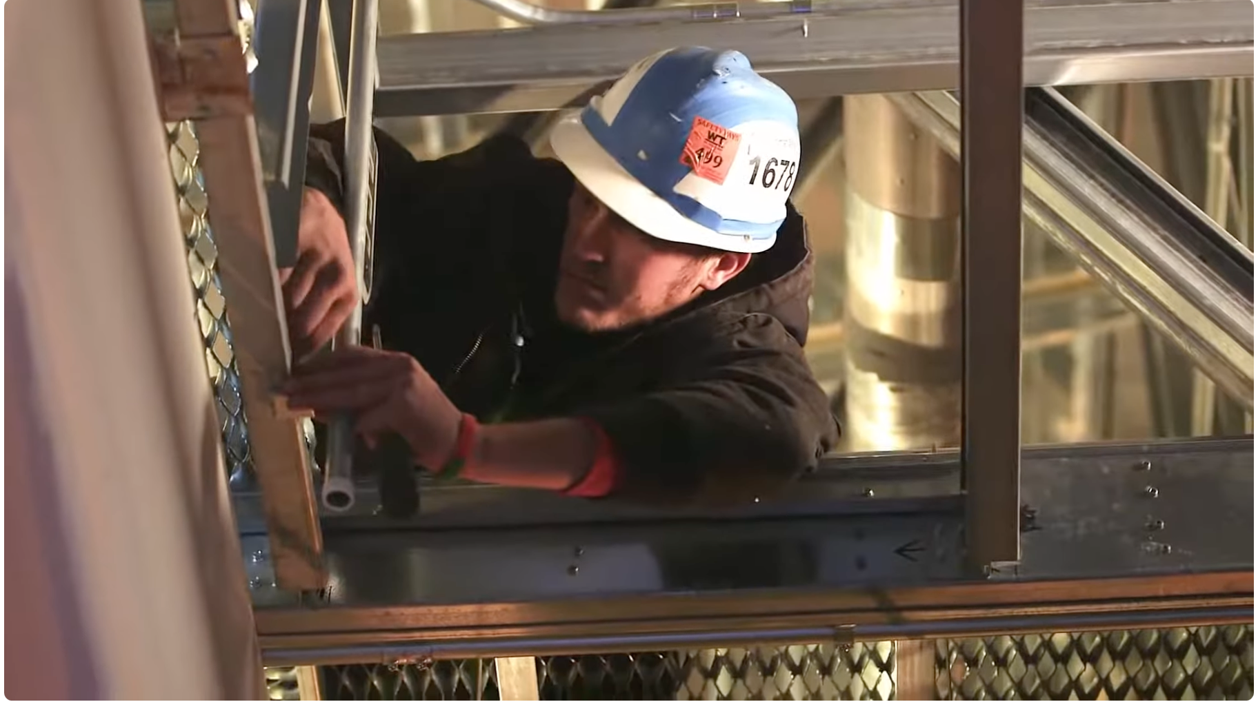A construction worker wearing a safety helmet and black jacket is installing or inspecting electrical components inside a metallic structure at a construction site.