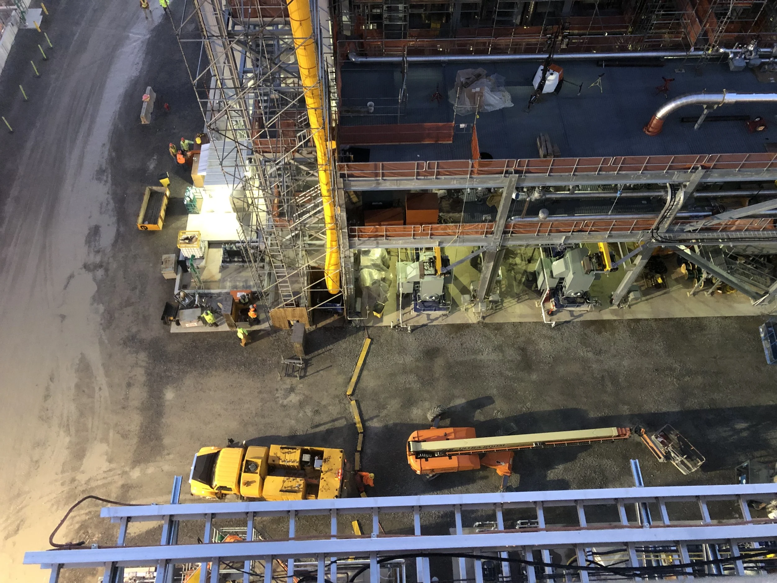 An aerial view of a construction site with workers, equipment, and scaffolding. There are vehicles, large pipes, and construction materials visible.